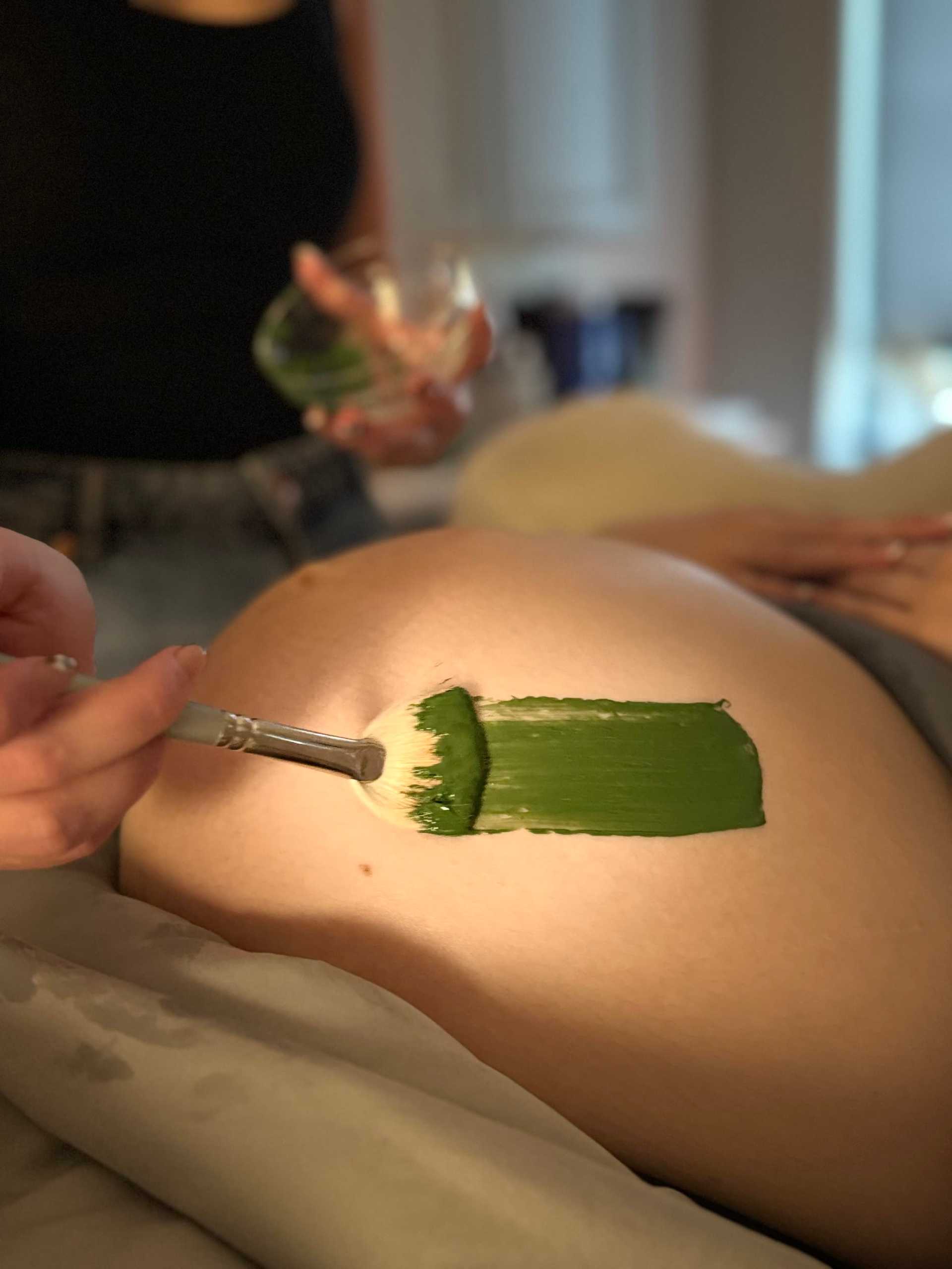 Person painting a green stripe on a pregnant belly with a brush, holding a small jar.