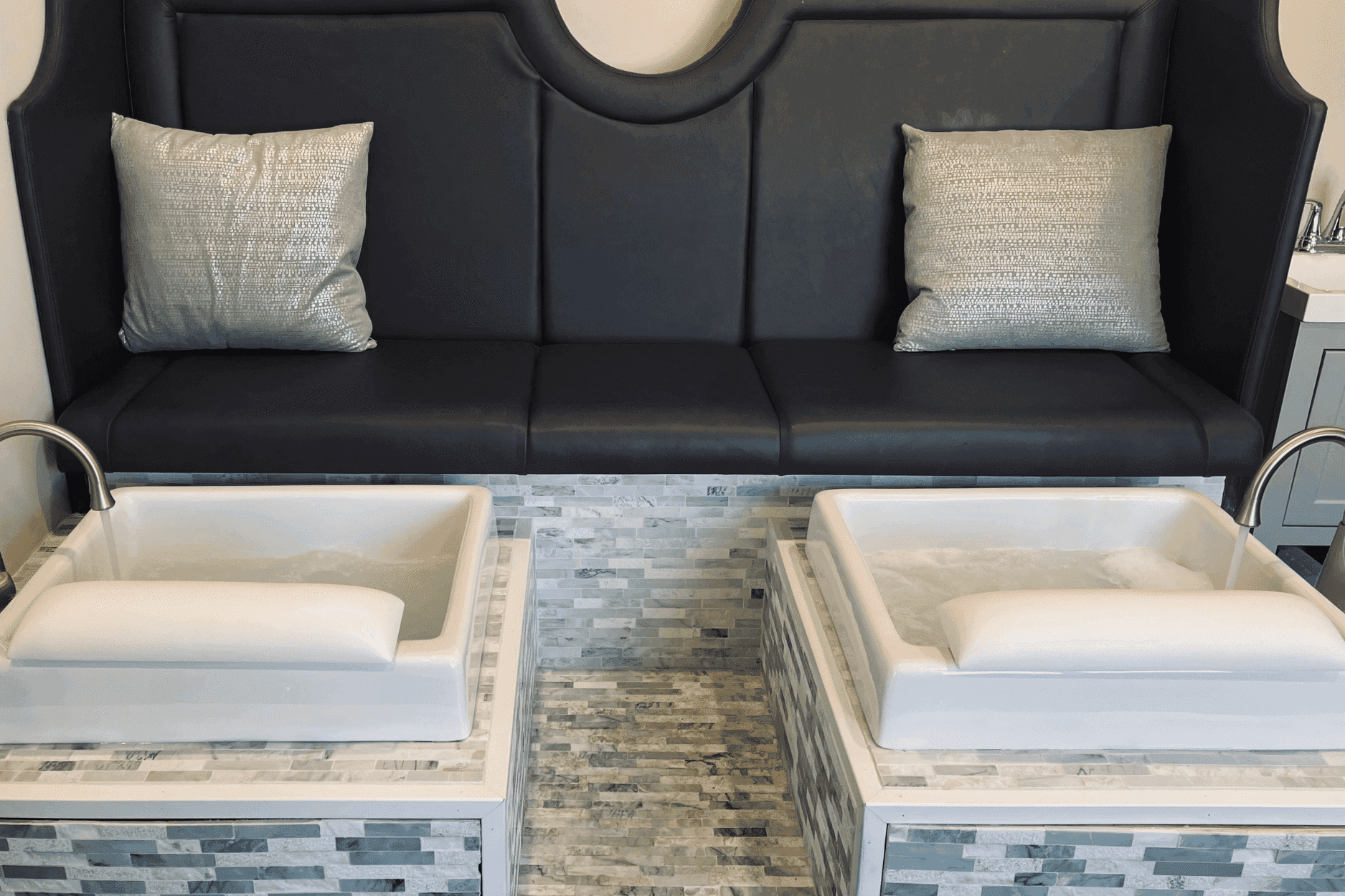 Modern pedicure station with cushioned bench and two foot spas in a sleek, tiled setting.
