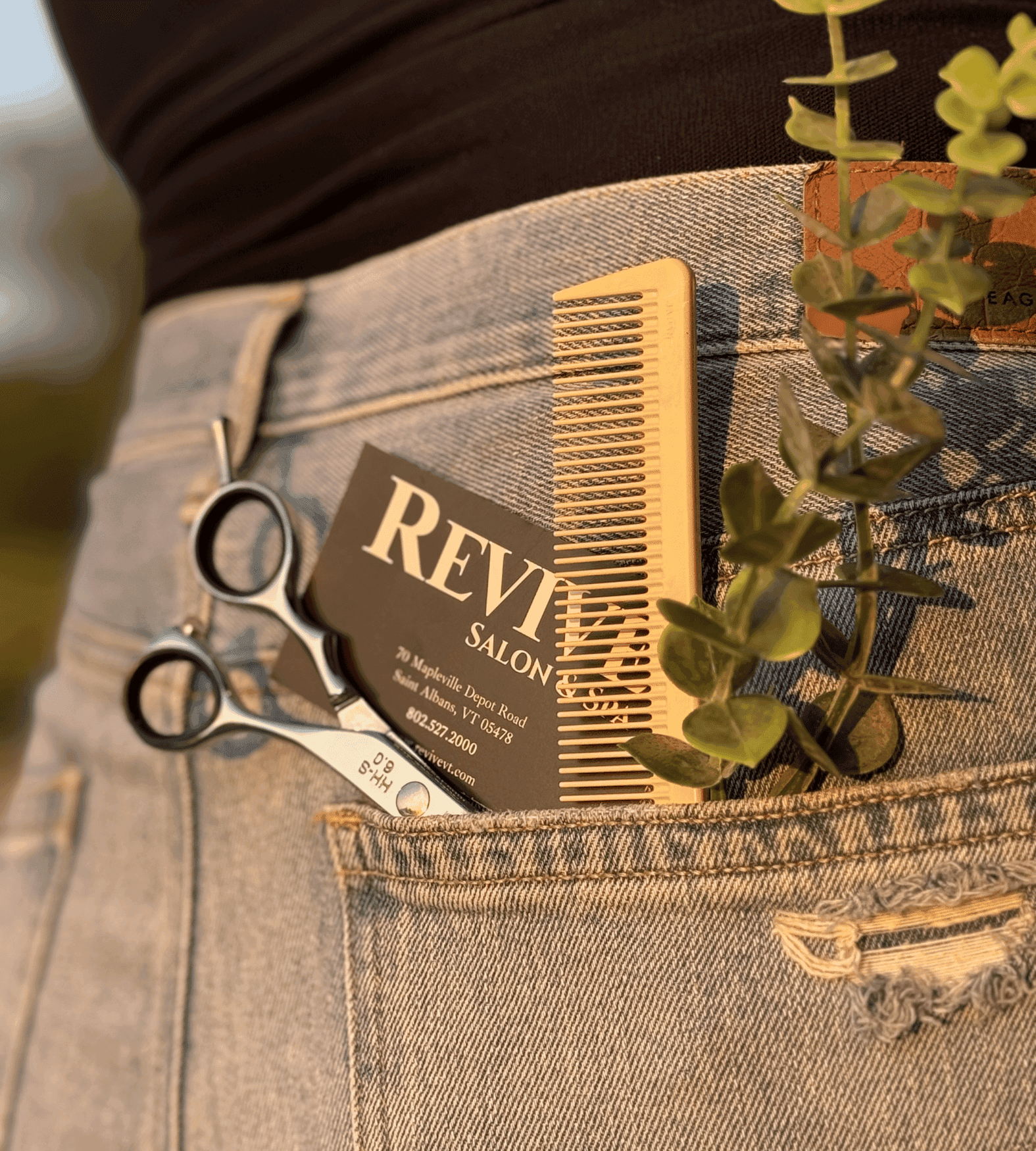 Scissors, comb, business card, and plant in back pocket of jeans in golden-hour lighting.